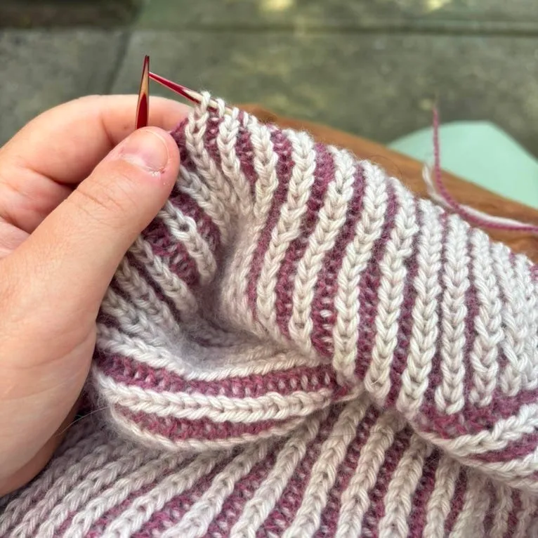 A hand holding knitting needles over a work in progress of two-color brioche ribbing in cream and dusty plum.